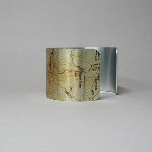 Puede incluir: Un brazalete de metal plateado con un mapa impreso de una regi&oacute;n monta&ntilde;osa. El mapa muestra la ubicaci&oacute;n de "Typooh Crater" y "Scott Pass".