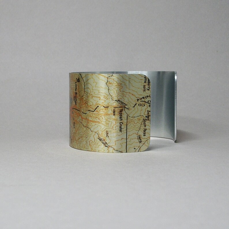 Puede incluir: Un brazalete de metal plateado con un mapa impreso de una regi&oacute;n monta&ntilde;osa. El mapa muestra la ubicaci&oacute;n de "Typooh Crater" y "Scott Pass".