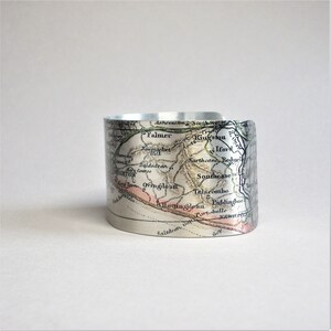 May include: A silver metal cuff bracelet featuring a vintage map of the Brighton area of England printed on it. The map shows the names of towns and villages, including Falmer, King'ston, Ifor, Southease, Roduel, Baldadean, Brighton, Ovingdean, Telscombe, Rottingdean, Liddinghoe, Saltdean, and Portobello.