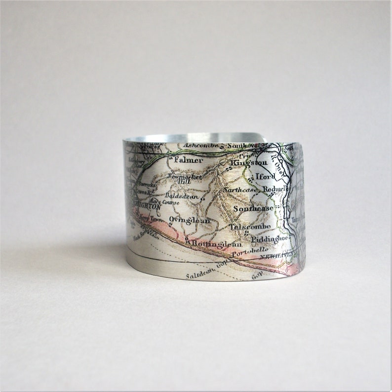 May include: A silver metal cuff bracelet featuring a vintage map of the Brighton area of England printed on it. The map shows the names of towns and villages, including Falmer, King'ston, Ifor, Southease, Roduel, Baldadean, Brighton, Ovingdean, Telscombe, Rottingdean, Liddinghoe, Saltdean, and Portobello.