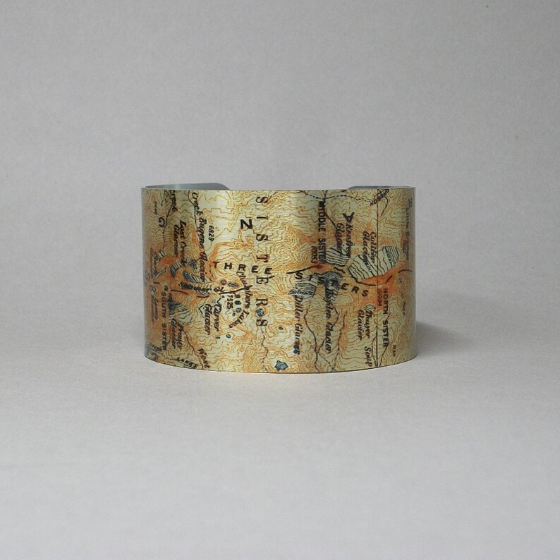 Puede incluir: Un brazalete de metal con un mapa de la Wilderness de las Tres Hermanas en Oreg&oacute;n. El mapa est&aacute; impreso en marr&oacute;n y dorado sobre un fondo plateado. El texto del mapa incluye "Three Sisters", "Middle Sister", "South Sister" y "Glacier".