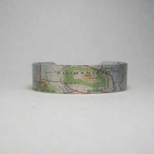 May include: A silver metal cuff bracelet with a map of Mount Kilimanjaro printed on it. The map shows the location of the mountain, the surrounding area, and the towns of Arusha and Moshi.