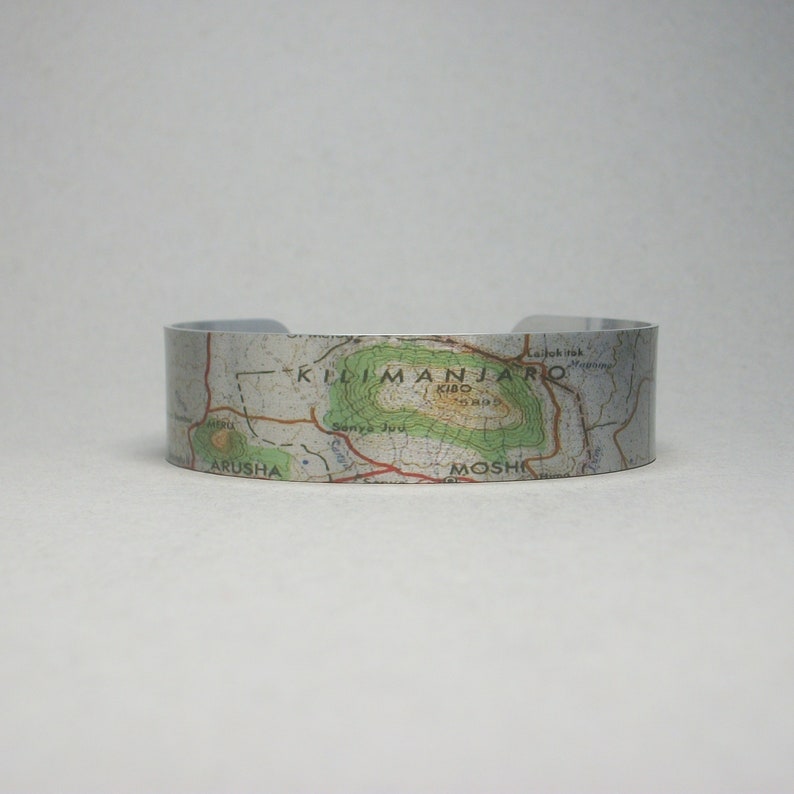 May include: A silver metal cuff bracelet with a map of Mount Kilimanjaro printed on it. The map shows the location of the mountain, the surrounding area, and the towns of Arusha and Moshi.