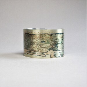 May include: A silver cuff bracelet with a vintage map of a coastal area printed on it. The map is in shades of green, brown, and blue.