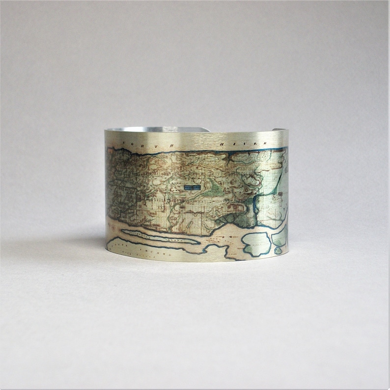 May include: A silver cuff bracelet with a vintage map of a coastal area printed on it. The map is in shades of green, brown, and blue.