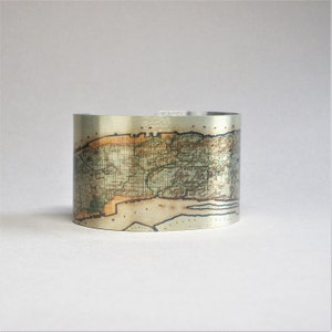 May include: A silver metal cuff bracelet with a vintage map of New York City printed on it. The map is in shades of brown, green, and blue.