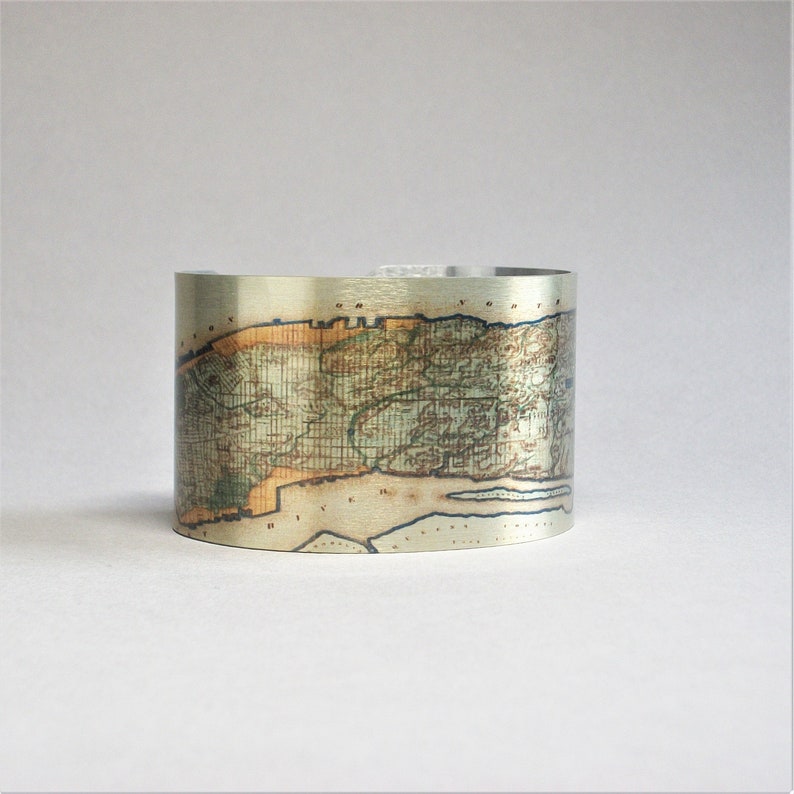 May include: A silver metal cuff bracelet with a vintage map of New York City printed on it. The map is in shades of brown, green, and blue.