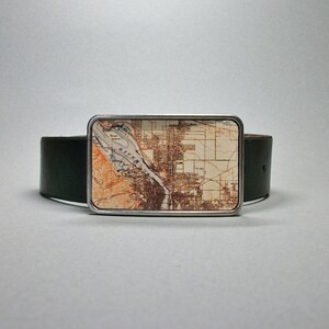 May include: A silver belt buckle featuring a vintage map of a city printed on it. The map is in shades of brown, beige, and white. The buckle is attached to a dark green leather belt.