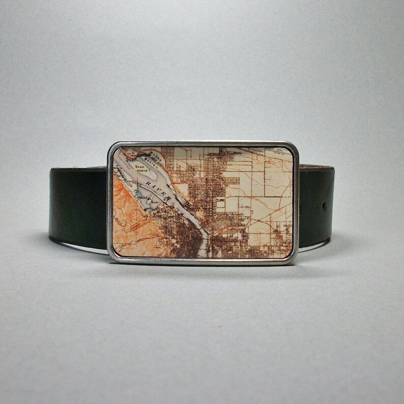 May include: A silver belt buckle featuring a vintage map of a city printed on it. The map is in shades of brown, beige, and white. The buckle is attached to a dark green leather belt.