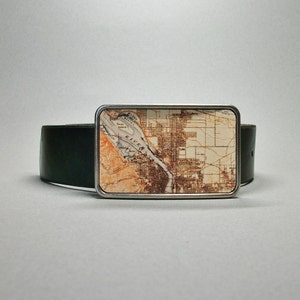 May include: A silver belt buckle featuring a vintage map design of a city and river. The map is printed on a cream background with brown and orange accents. The buckle is attached to a black leather belt.