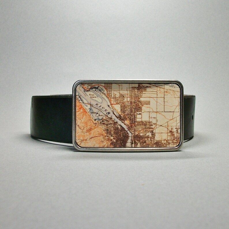 May include: A silver belt buckle featuring a vintage map design of a city and river. The map is printed on a cream background with brown and orange accents. The buckle is attached to a black leather belt.