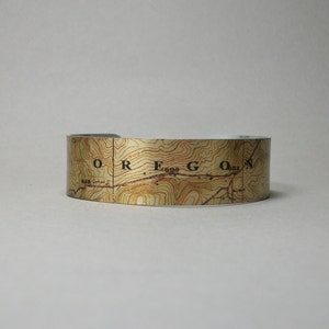 May include: A wide, open cuff bracelet featuring a topographic map design. The map is printed in shades of brown and tan, with the word "OREGON" visible. The bracelet is made of a shiny, metallic material.