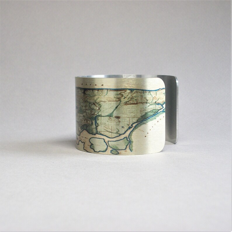 May include: A silver metal cuff bracelet with a vintage map design printed on the front. The map is in shades of green, brown, and blue.