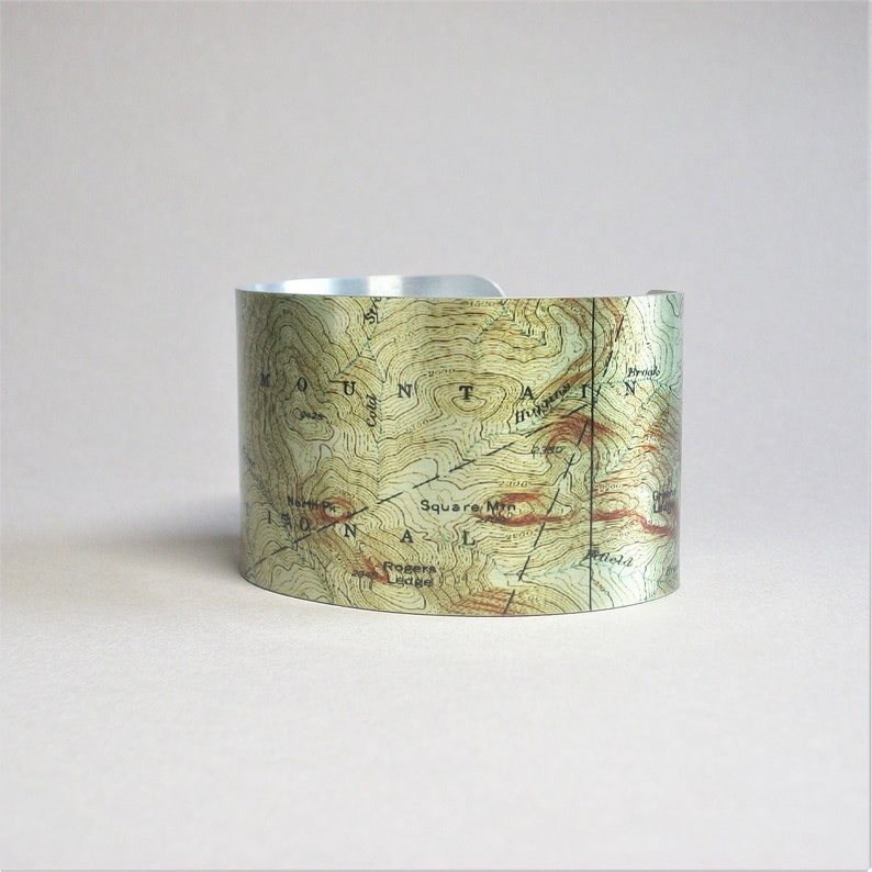 Pu&ograve; includere: Un braccialetto rigido in metallo argentato con un design a mappa topografica. La mappa mostra l'area intorno al Monte Washington, nel New Hampshire, con il testo "Mount Washington" e "Square Mtn" visibile.