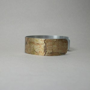 May include: A silver-toned metal cuff bracelet with a topographic map design. The map features brown contour lines and text, including "Pacific" and "Jones Creek", set against a beige background. The bracelet has a smooth, polished finish.