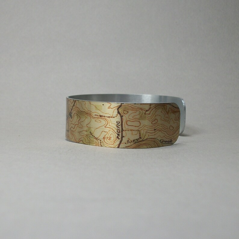 May include: A silver-toned metal cuff bracelet with a topographic map design. The map features brown contour lines and text, including "Pacific" and "Jones Creek", set against a beige background. The bracelet has a smooth, polished finish.