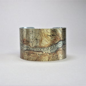 May include: A silver metal cuff bracelet with a vintage map design. The map features the Columbia River and surrounding area, with details like rivers, mountains, and roads. The map is printed in brown and gold tones on a silver background.