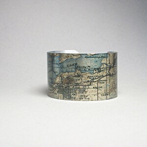 May include: A wide, silver cuff bracelet featuring a vintage map design in shades of blue, brown, and white. The map details include geographical features and place names. The bracelet has a slightly distressed appearance.
