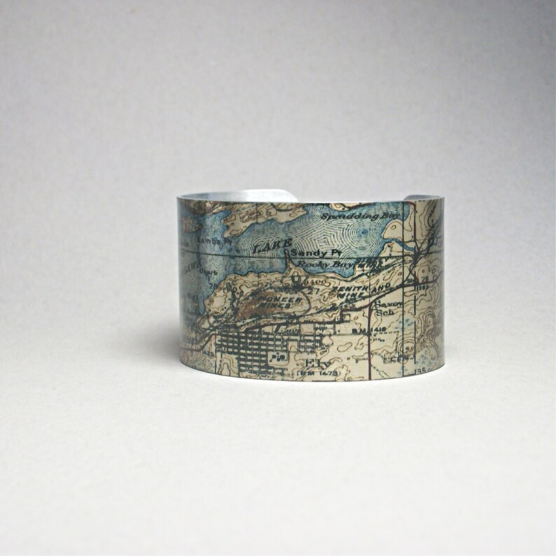 May include: A wide, silver cuff bracelet featuring a vintage map design in shades of blue, brown, and white. The map details include geographical features and place names. The bracelet has a slightly distressed appearance.