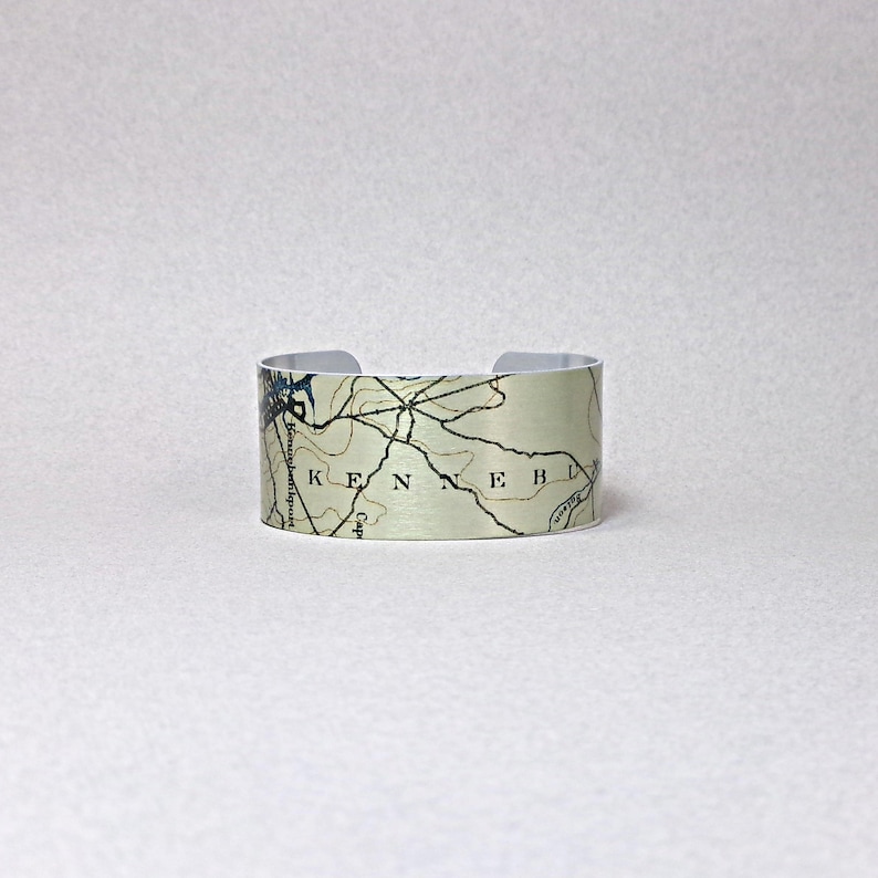 May include: A wide, silver-toned cuff bracelet featuring a map design. The map has black lines and the word "KENNEB" is visible. The bracelet is open at the back and has a neutral background.