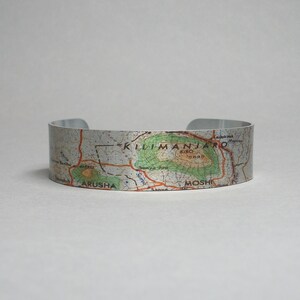 May include: A silver metal bracelet with a map of Mount Kilimanjaro printed on it. The map shows the location of the summit, Kibo, and the towns of Moshi and Arusha.