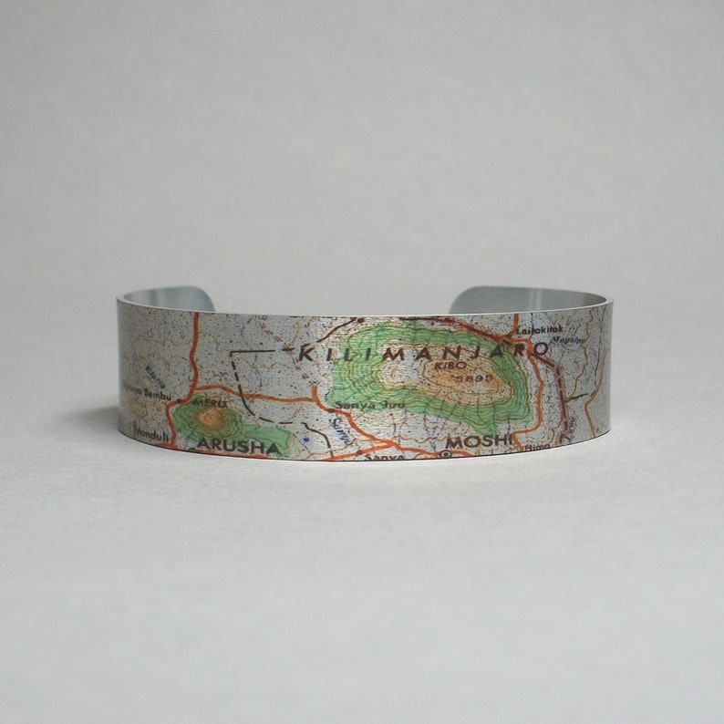 May include: A silver metal bracelet with a map of Mount Kilimanjaro printed on it. The map shows the location of the summit, Kibo, and the towns of Moshi and Arusha.