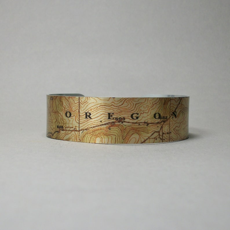 May include: A wide, open cuff bracelet featuring a topographic map design. The map is printed in shades of brown and tan, with the word "OREGON" visible. The bracelet is a neutral accessory.