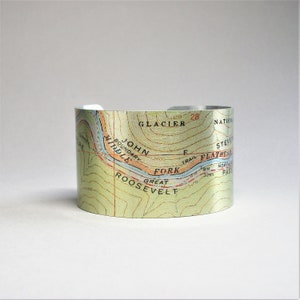 May include: A metal cuff bracelet with a map of Glacier National Park printed on it. The map shows the John Boundary, Middle Fork, and Great Roosevelt areas. The text "GLACIER NATIONAL PARK" is visible on the map.