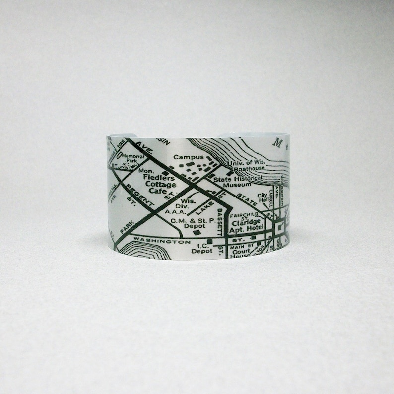 May include: A silver-toned metal cuff bracelet featuring a black printed map design. The map displays street names like Regent St and Washington, along with landmarks such as the State Historical Museum, City Hall, and the University of Wisconsin Boathouse.