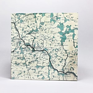 May include: A square map print featuring a vintage-style cartographic design of Algonquin Park. The map displays various lakes in a muted teal color against a cream background, with black lines representing railways and text labels for locations.
