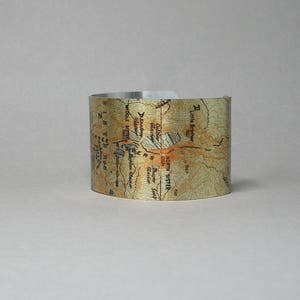 Puede incluir: Un brazalete de metal plateado con un dise&ntilde;o de mapa topogr&aacute;fico de las monta&ntilde;as North Sister y Middle Sister en Oreg&oacute;n. El mapa incluye los nombres de las monta&ntilde;as, los glaciares y otras caracter&iacute;sticas geogr&aacute;ficas.