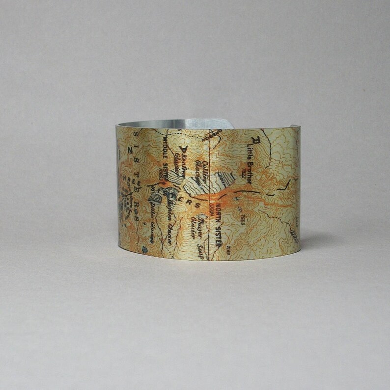 Puede incluir: Un brazalete de metal plateado con un dise&ntilde;o de mapa topogr&aacute;fico de las monta&ntilde;as North Sister y Middle Sister en Oreg&oacute;n. El mapa incluye los nombres de las monta&ntilde;as, los glaciares y otras caracter&iacute;sticas geogr&aacute;ficas.