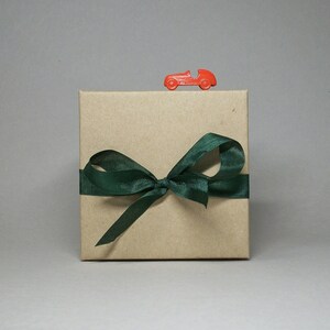 May include: A square, kraft paper gift box tied with a dark green satin ribbon and bow. A small, red toy race car sits on top of the box. The background is a solid, neutral gray.