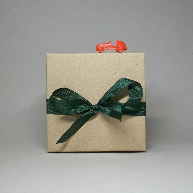 May include: A square, kraft paper gift box tied with a dark green satin ribbon and bow. A small, red toy race car sits on top of the box. The background is a solid, neutral gray.