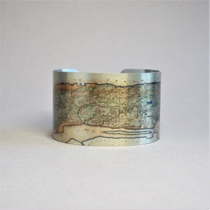 May include: A silver metal cuff bracelet with an antique map of Long Island, New York printed on it. The map is in shades of brown, green, and blue.