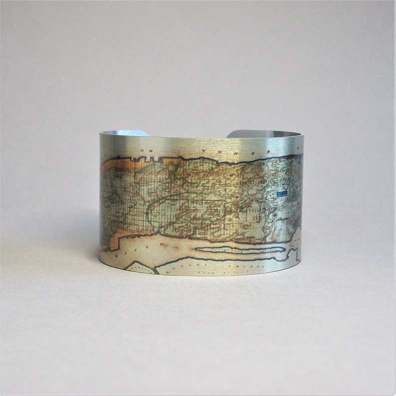 May include: A silver metal cuff bracelet with an antique map of Long Island, New York printed on it. The map is in shades of brown, green, and blue.