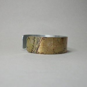May include: A silver-colored cuff bracelet with a topographic map design. The map features shades of tan, brown, and black, with visible contour lines and text. The bracelet is made of a shiny metal and has a simple, modern design.