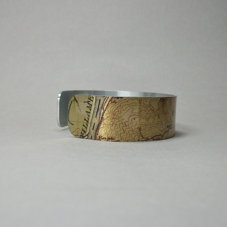 May include: A silver-colored cuff bracelet with a topographic map design. The map features shades of tan, brown, and black, with visible contour lines and text. The bracelet is made of a shiny metal and has a simple, modern design.