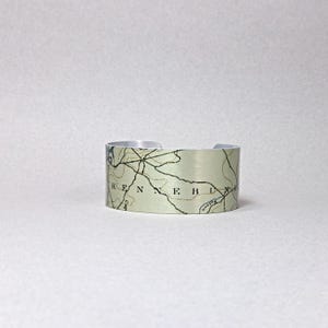 May include: Silver-toned metal cuff bracelet featuring a map design. The map includes the word "KENNEB" and other geographical details. The bracelet has a brushed metal finish and a simple, open design.