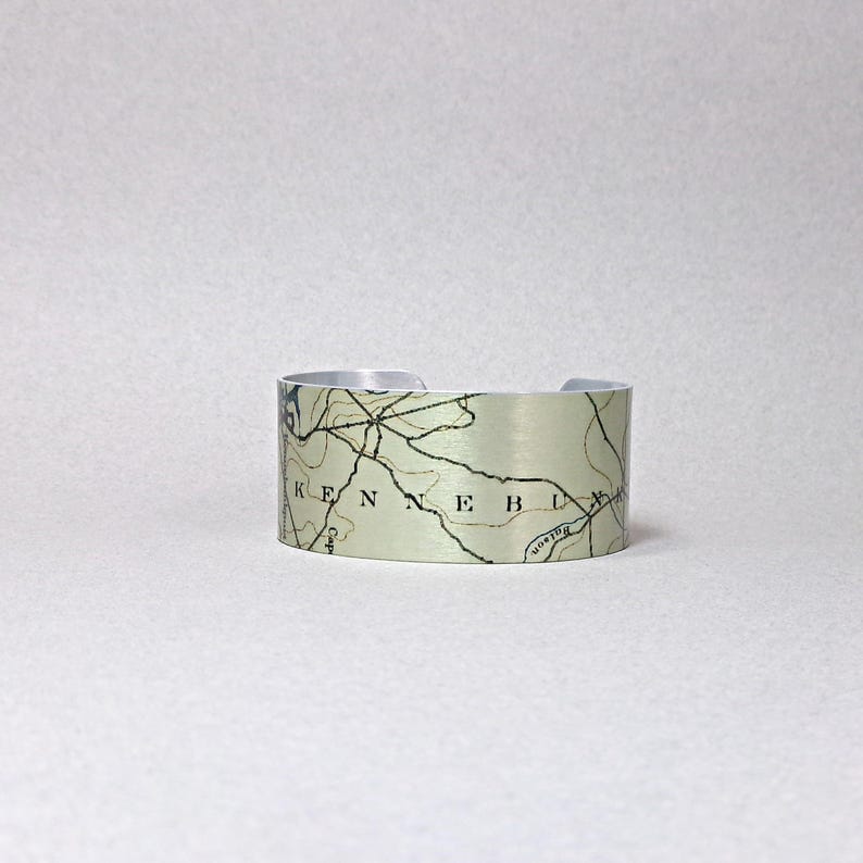 May include: Silver-toned metal cuff bracelet featuring a map design. The map includes the word "KENNEB" and other geographical details. The bracelet has a brushed metal finish and a simple, open design.