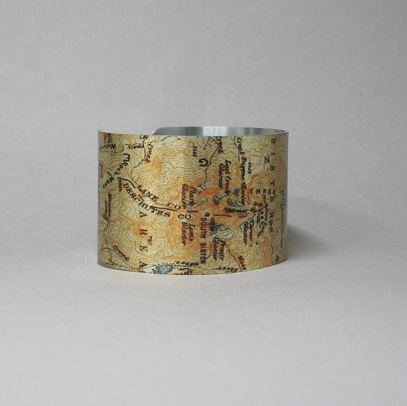 Puede incluir: Un brazalete de metal plateado con un dise&ntilde;o de mapa vintage que incluye el texto "South Sister", "Lane Co", "Deschutes Co", "Lost Creek Glacier", "Fronty Glacier" y "Three Sisters".