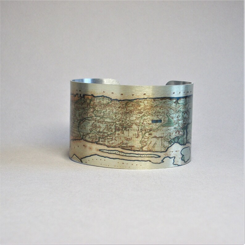 May include: A silver metal cuff bracelet with a vintage map design. The map is printed on the metal and shows a detailed view of a landmass with rivers and other geographical features.