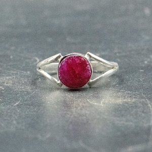 Op de afbeelding: Een zilveren ring met een ronde, felroze edelsteen in het midden. De ring heeft een eenvoudig, elegant ontwerp met twee delicate punten aan weerszijden van de steen.