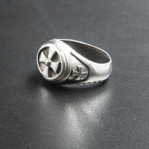 Könnte beinhalten: Ein silberner Ring mit einem Kreuzdesign. Der Ring liegt auf einer schwarzen Oberfläche.