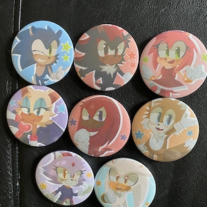 May include: A collection of eight round pinback buttons featuring various characters from the Sonic the Hedgehog video game series. Each button has a different character design, with vibrant colors and star accents. The buttons are arranged on a dark surface.