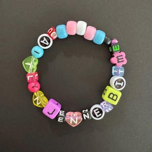 May include: A colorful beaded bracelet with the name "JENNE" spelled out using various letter beads. The bracelet includes beads in shades of pink, blue, white, yellow, and green. The bracelet is on a dark gray surface.