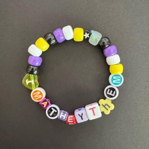 May include: A colourful beaded bracelet featuring a mix of beads in white, yellow, purple, and black. The bracelet spells out "THEY/THEM" with letter beads. Other beads are shaped like a star and a flower.