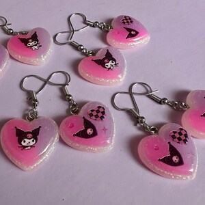 以下が含まれることがあります： ピンクと白のグリッターの縁取りが施されたハート型のイヤリング。各イヤリングには、白黒のチェック柄のハートまたはキャラクターイラストが描かれた漫画のキャラクターが描かれています。イヤリングにはシルバーカラーのフックが付いています。