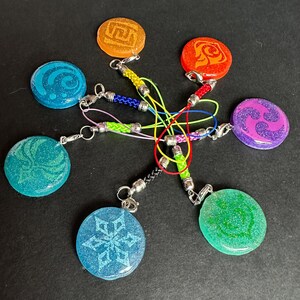 May include: A collection of seven colorful resin charms with silver-tone hardware. Each charm is round and features a different design in shades of blue, green, orange, red, and purple. The charms are connected by a network of colorful cords.