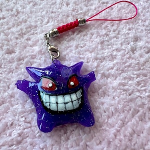 May include: A purple, glittery charm featuring a cartoon monster with a wide grin, red eyes, and white teeth. The charm has a red woven strap and a pink loop. The charm is likely a collectible or accessory.
